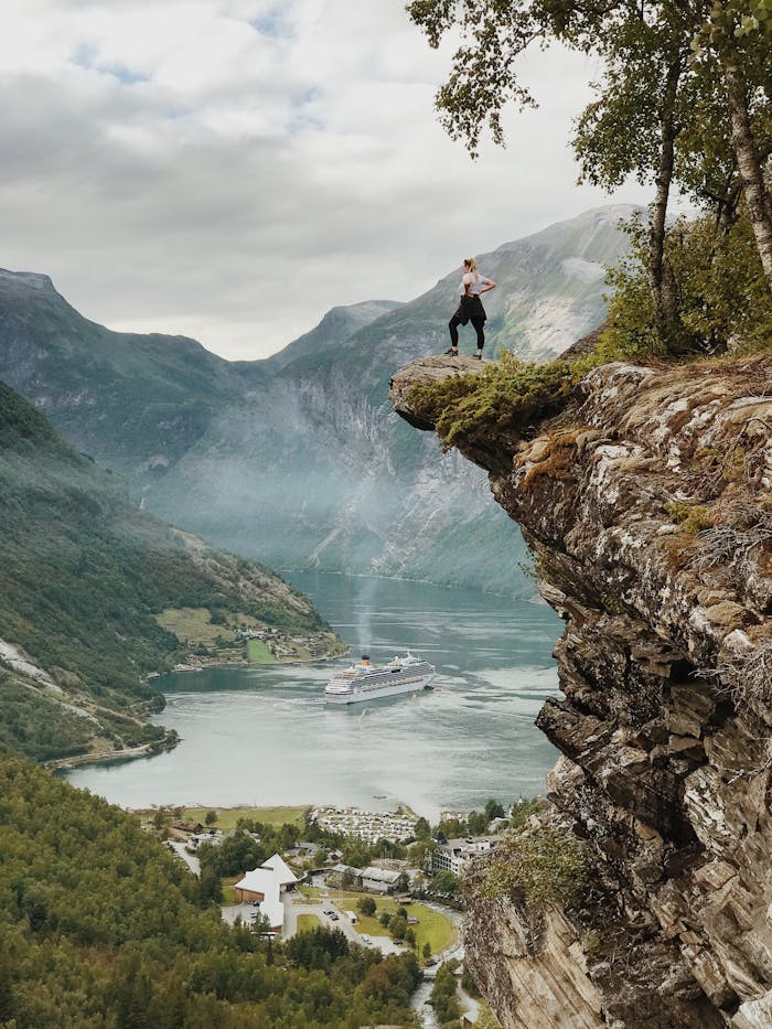 heros-img Person enjoys breathtaking view from a cliff above Geiranger Fjord, Norway, with clear skies and cruise ship below.